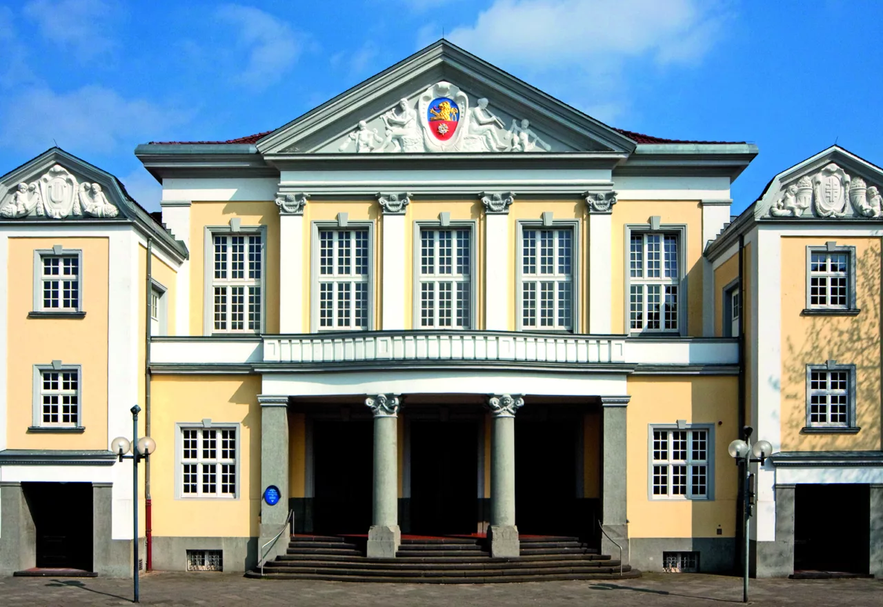 Das Bild zeigt die Außenansicht des Eingangs der Festhalle Viersen. Die Fassade zeigt klassizistische Formelemente wie Säulen, Dreiecksgiebel und Pilaster. Das Gebäude ist hellgelb, die Säulen und Ornamente sind weiß.