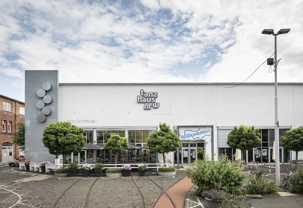 Das Foto zeigt eine Außenansicht des tanzhaus nrw mit dem Zugang über den Hof, über den die alten Schienen verlaufen, sowie den Gastronomie-Außenbereich. An der Fassade hängt der Schriftzug "tanzhaus nrw".