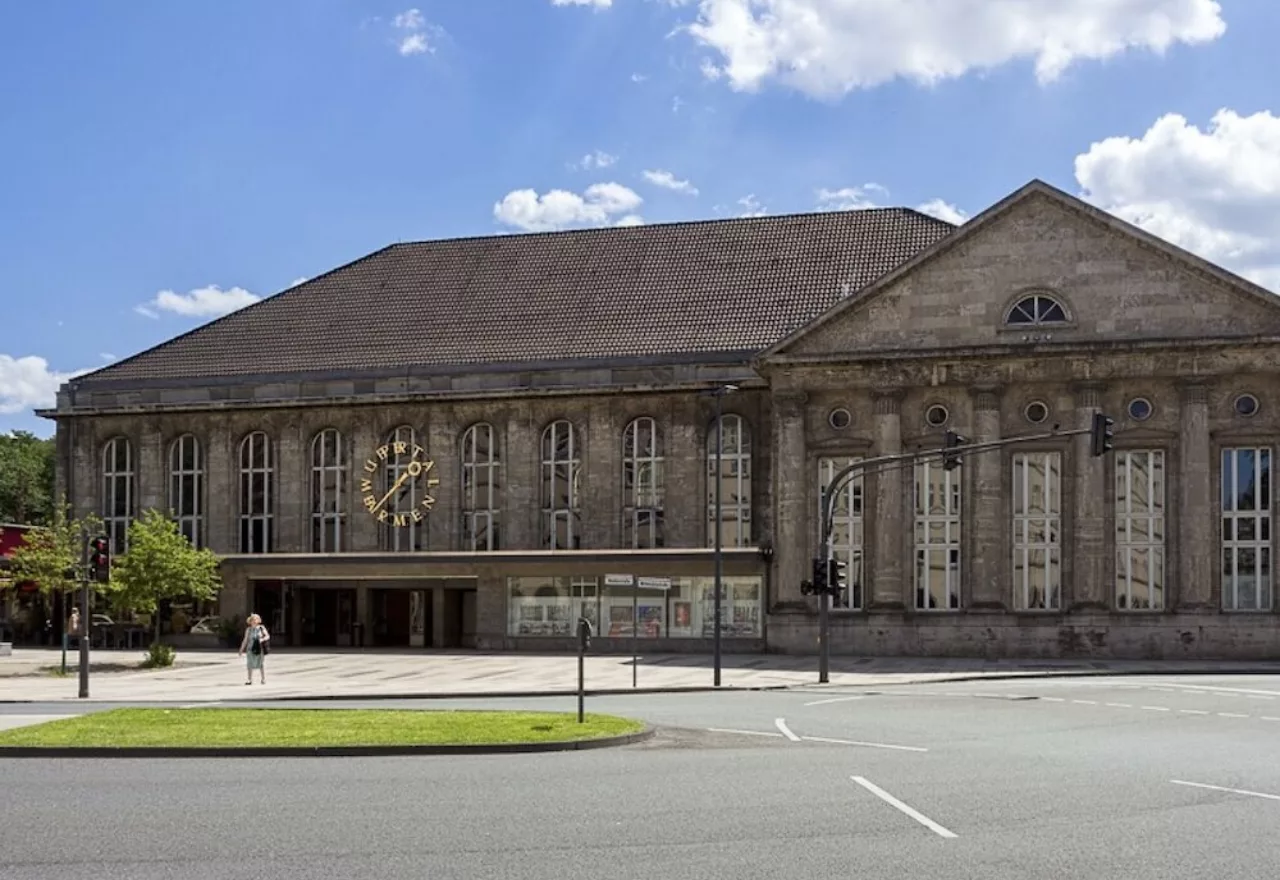 Das Bild zeigt das alte Bahnhofsgebäude in Wuppertal Barmen. Das Ziffernblatt der großen goldenen Uhr besteht aus den Buchstaben von "WUPPERTAL BARMEN".