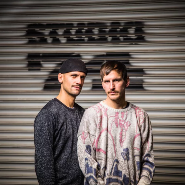 Das Bild zeigt links Daniel Ernesto Mueller und rechts Simon Hartmann, die vor einem beigen Rolltor stehen mit einem schwarzen Graffitti im Hintergrund.